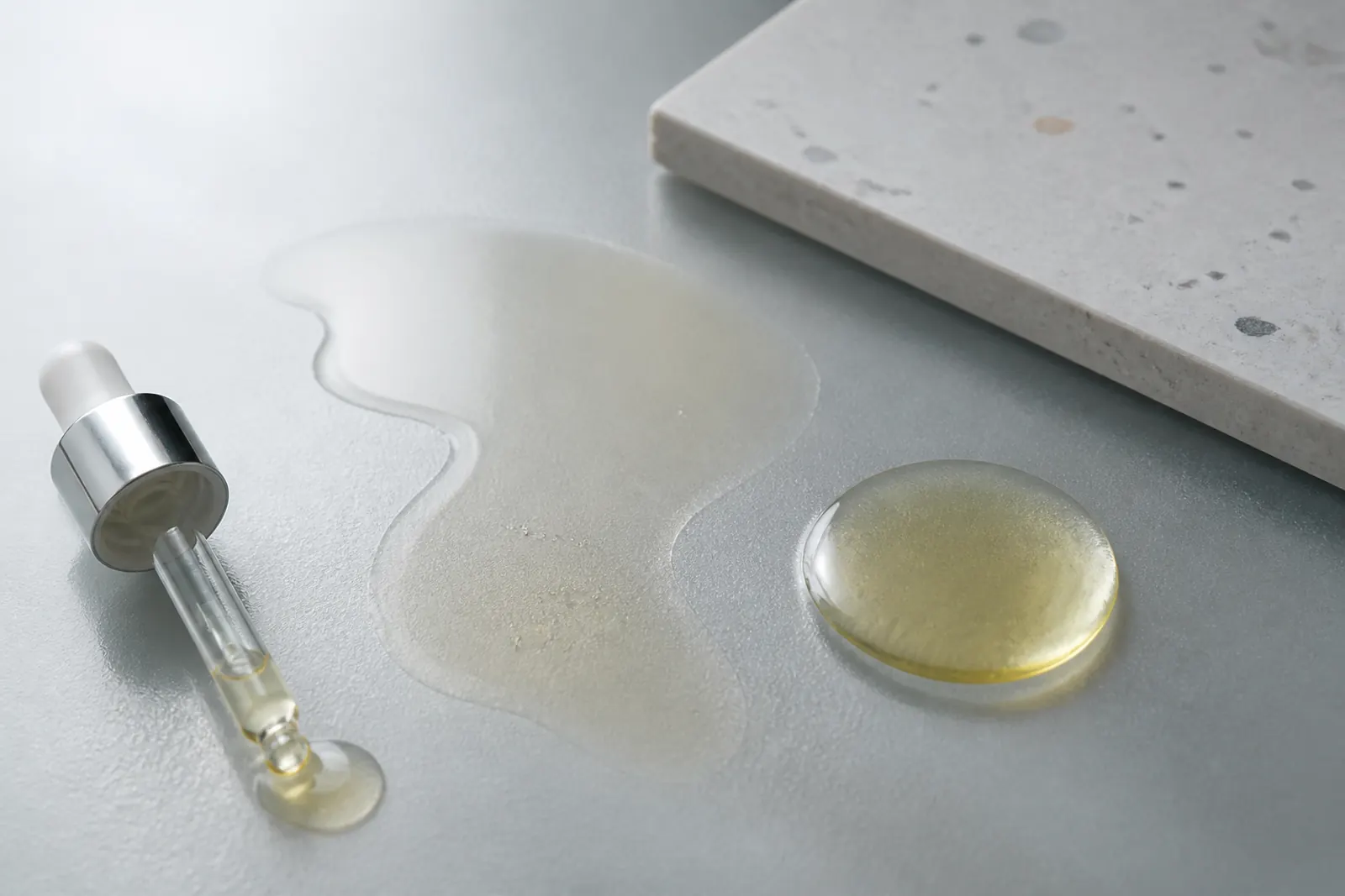 Two oil films on frosted glass, one thin and fast-spreading, one cushioned droplet, dropper and ceramic tile, no labels